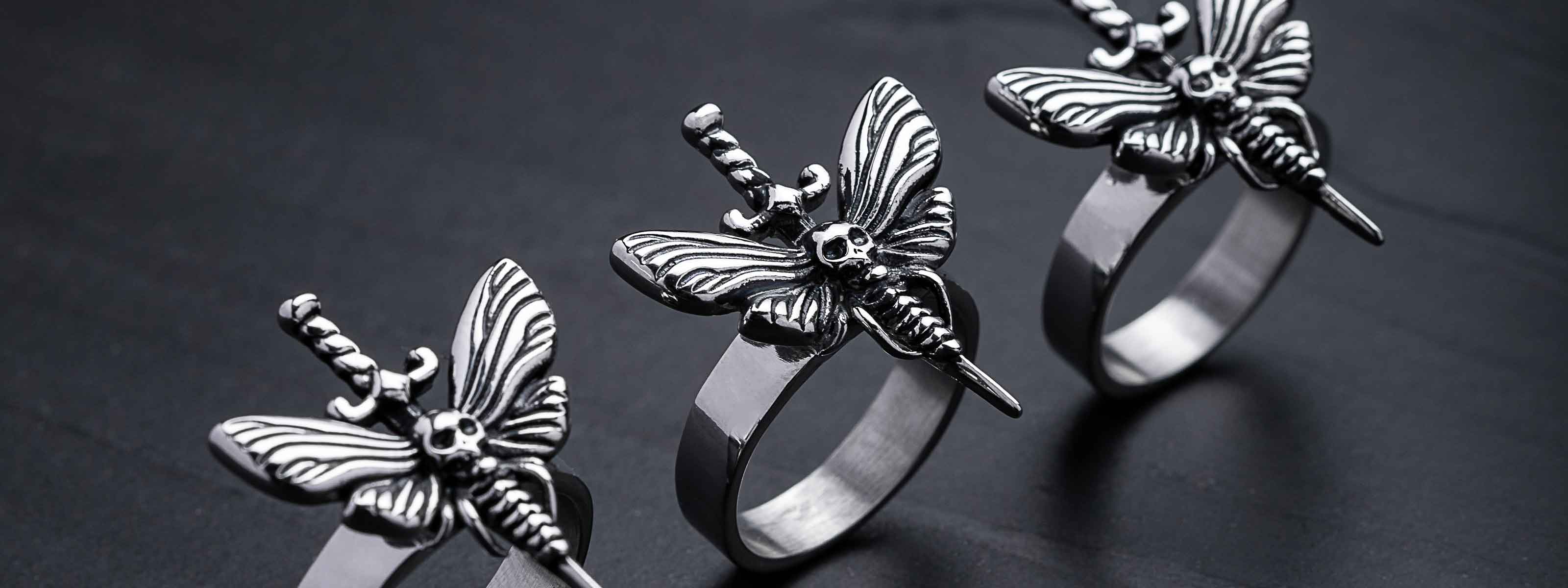 Three silver rings with butterfly designs, each featuring a sword and a skull detail in the center, displayed on a dark, matte surface.