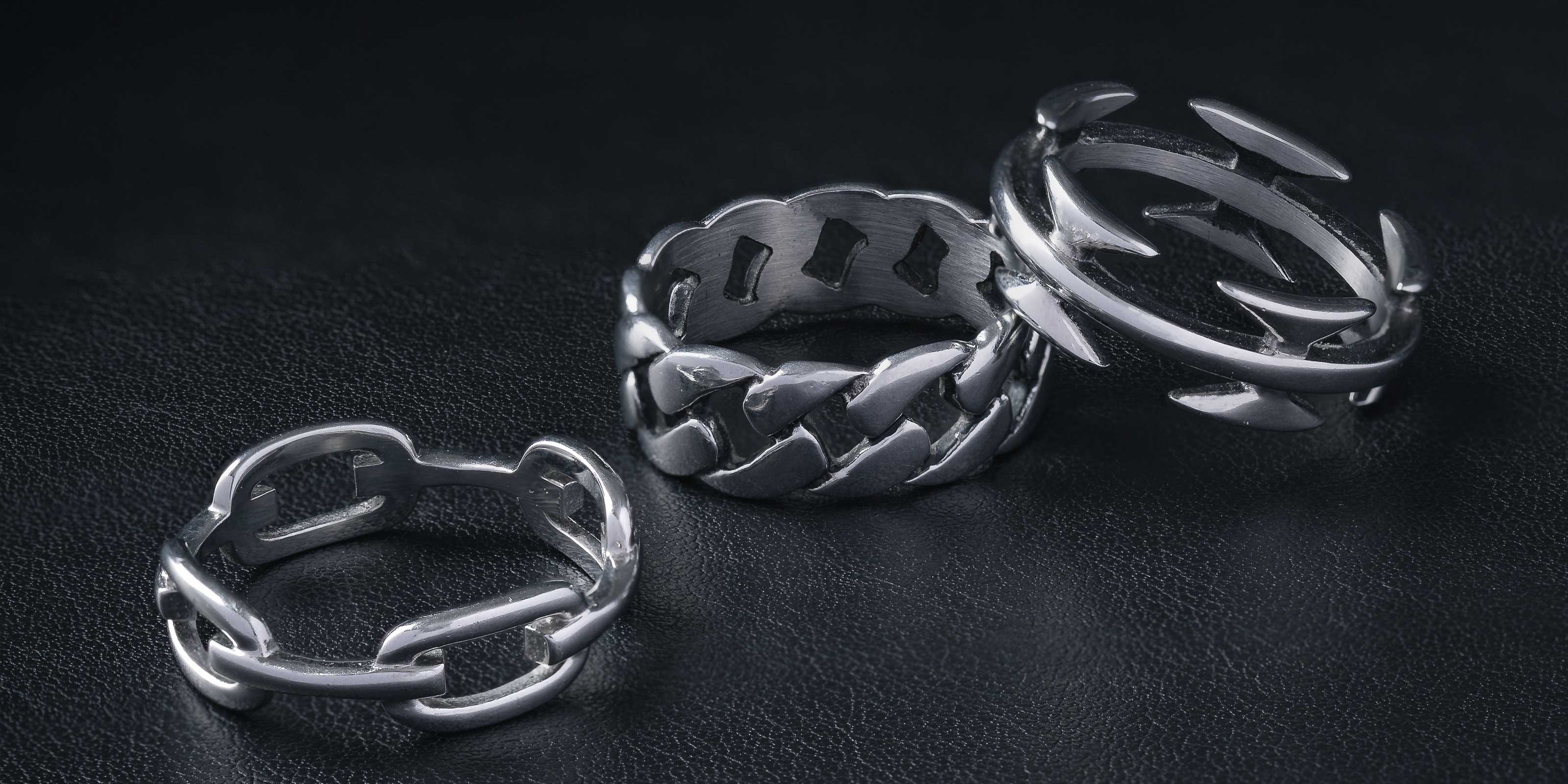 Three silver rings with bold, chunky chain-link designs are displayed on a dark, textured surface. Each ring features a different chain style and metallic finish.