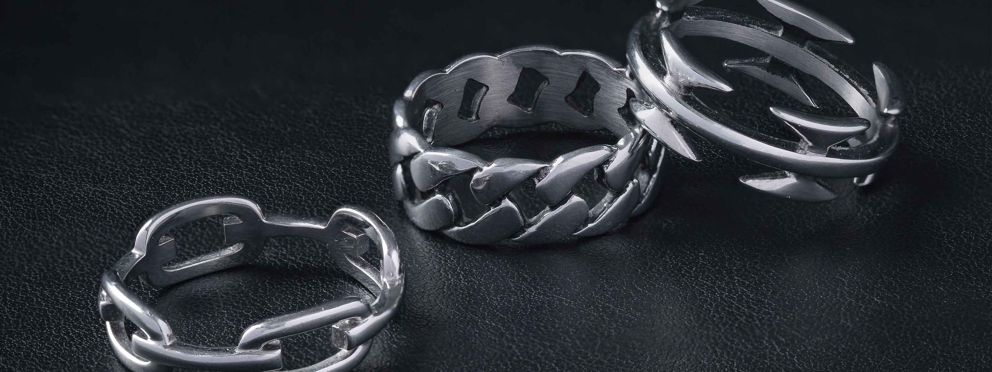 Three silver rings with bold, chain-link and geometric designs are displayed on a dark, textured surface.