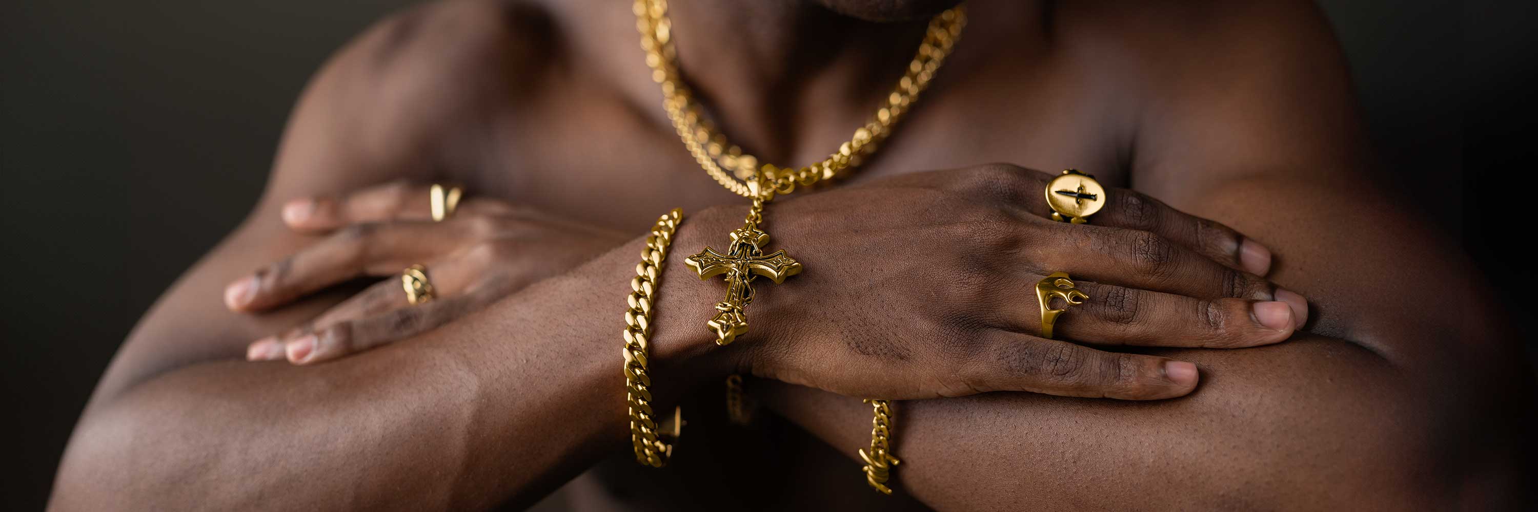 A shirtless person crosses their arms, displaying several gold chains, rings, and a cross pendant with a dark background.