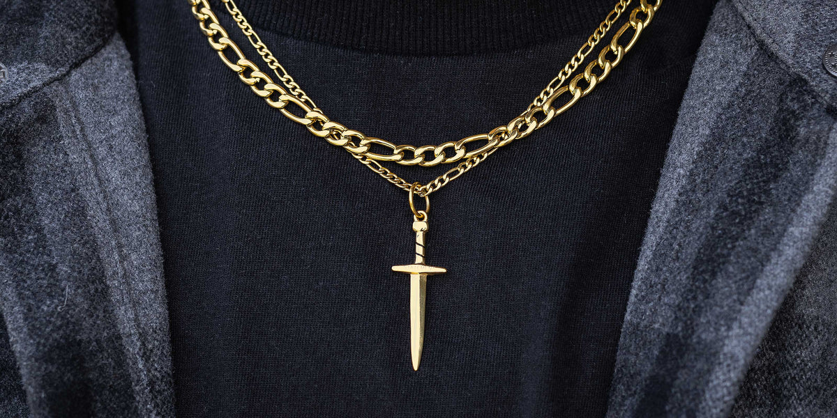 Two gold chains around a persons neck, one with a dagger pendant. The person is wearing a black shirt and a dark jacket.