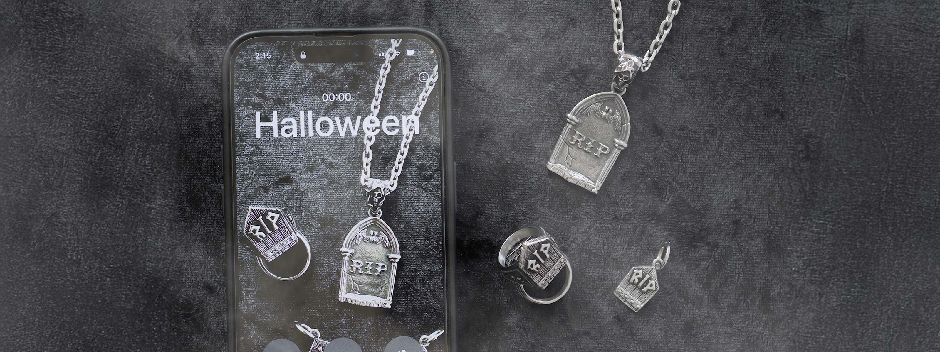 A phone displays Halloween alongside silver tombstone-shaped jewelry, including necklaces, rings, and pendants, all engraved with RIP, arranged on a dark, textured background.