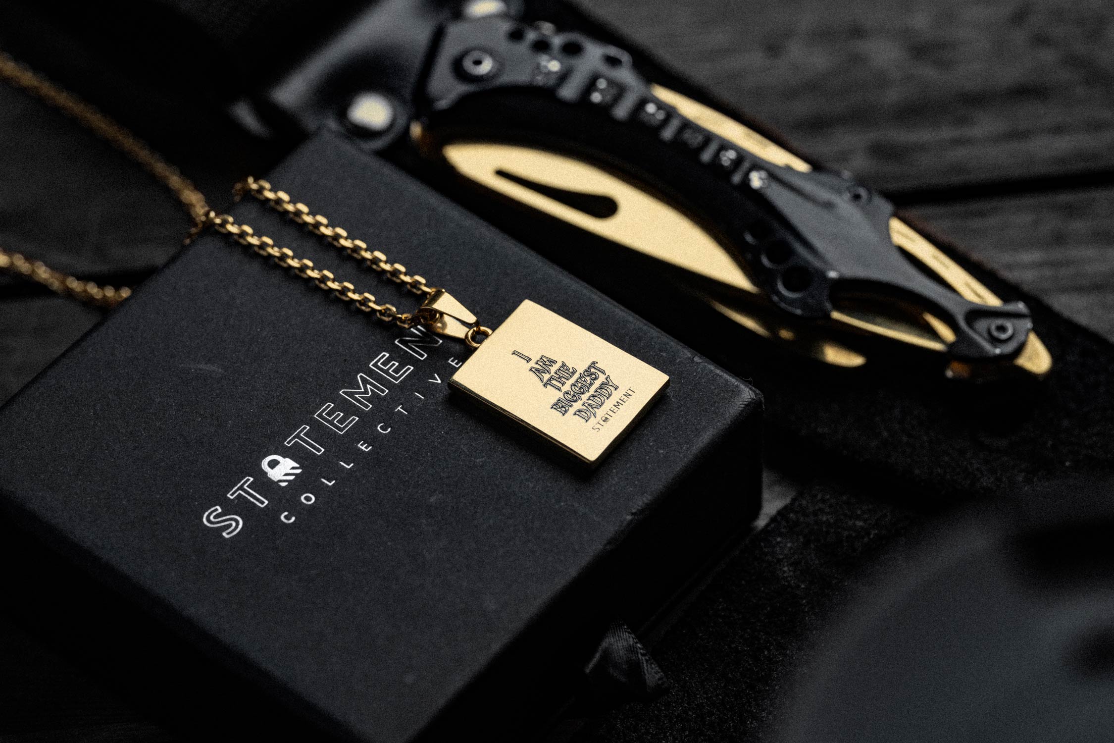A gold pendant necklace on a black box labeled “STATEMENT COLLECTIVE,” next to a black and gold folding knife on a dark wooden surface.