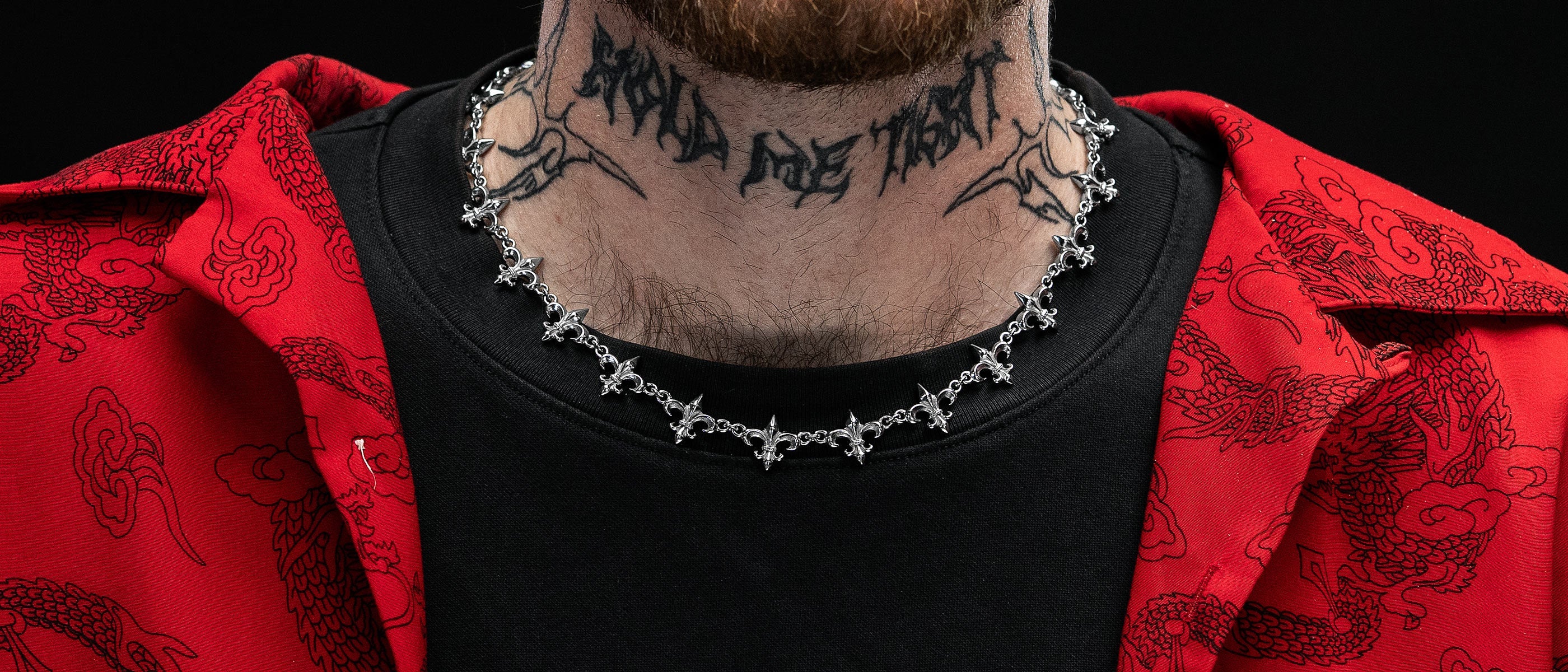 A person wearing a red patterned jacket, black shirt, a silver chain necklace with cross charms, and a neck tattoo that reads BIND ME THAT in black lettering.