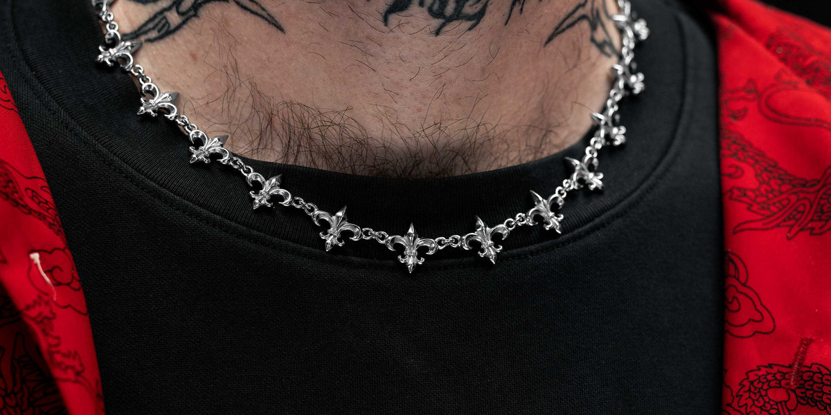 A person wearing a red patterned jacket, black shirt, a silver chain necklace with cross charms, and a neck tattoo that reads BIND ME THAT in black lettering.