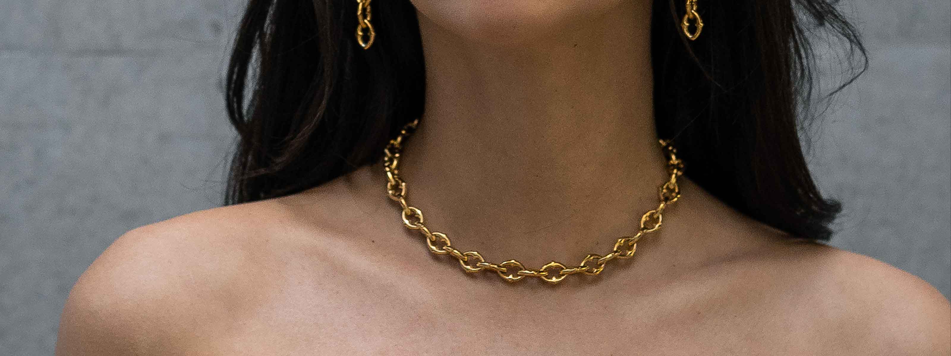 A woman wearing a gold chain necklace and matching gold chain earrings. Her shoulders are bare, and her dark hair is draped behind her shoulders against a neutral background.