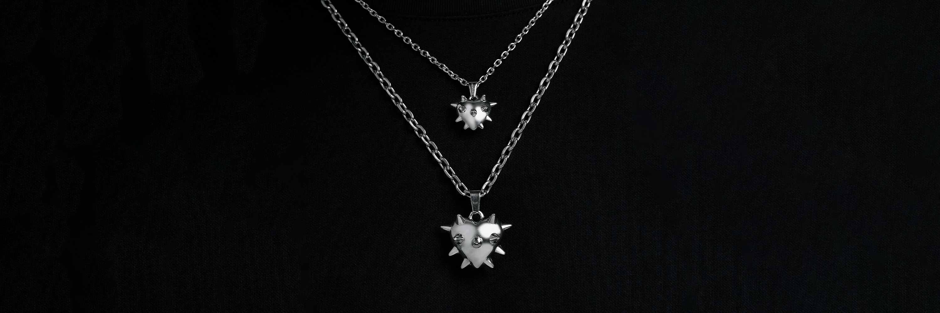 Two silver chain necklaces with spiked heart pendants are displayed against a black background, with one necklace shorter and the other longer.