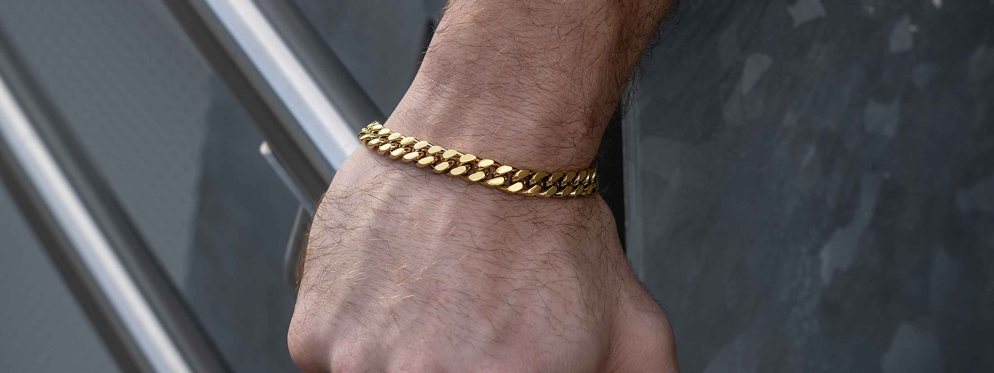 A close-up of a persons wrist wearing a thick gold chain bracelet against a dark, textured background with metal railings.