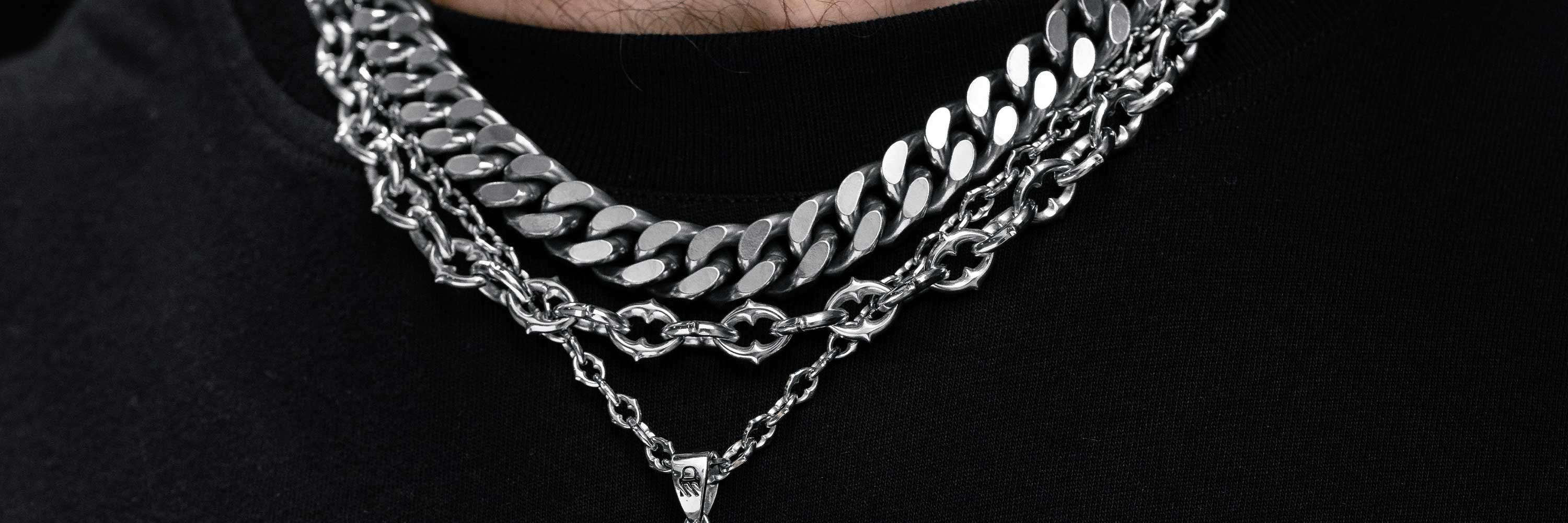 Close-up of a person wearing three silver chains of different styles and thicknesses over a black shirt; the lowest chain has a small pendant.