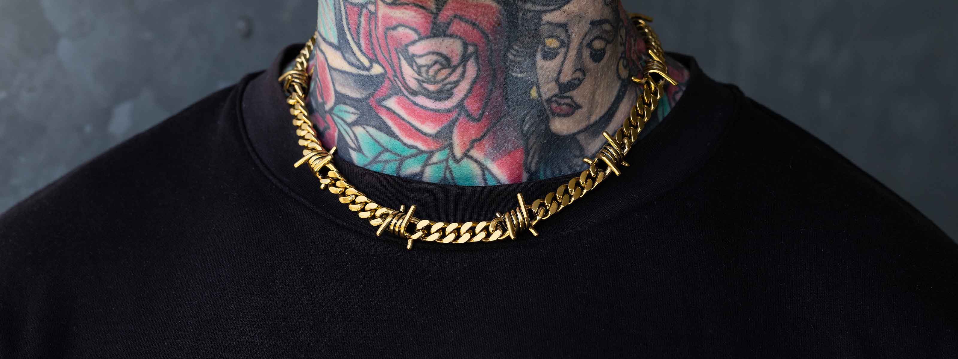 A person wearing a gold chain necklace with barbed wire details, dressed in a black shirt, showing colorful neck tattoos with floral and face designs.
