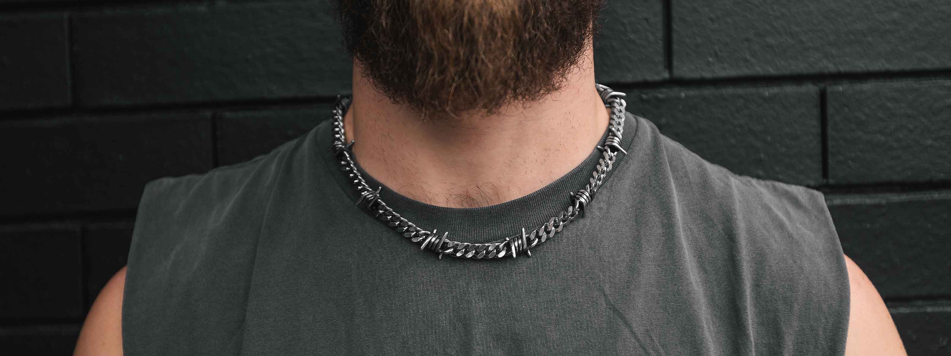 A close-up of a bearded person wearing a sleeveless gray shirt and a chunky silver chain necklace, standing in front of a black brick wall.