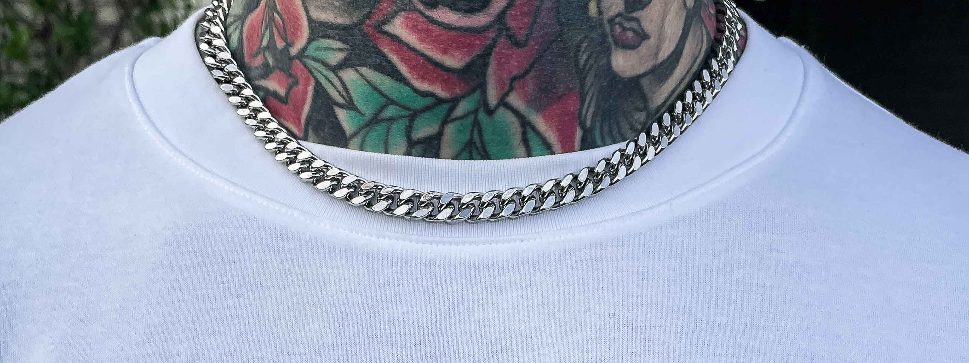 A person wearing a thick silver chain necklace over a white shirt, with colorful tattoo designs featuring a rose and a womans face visible on their neck.