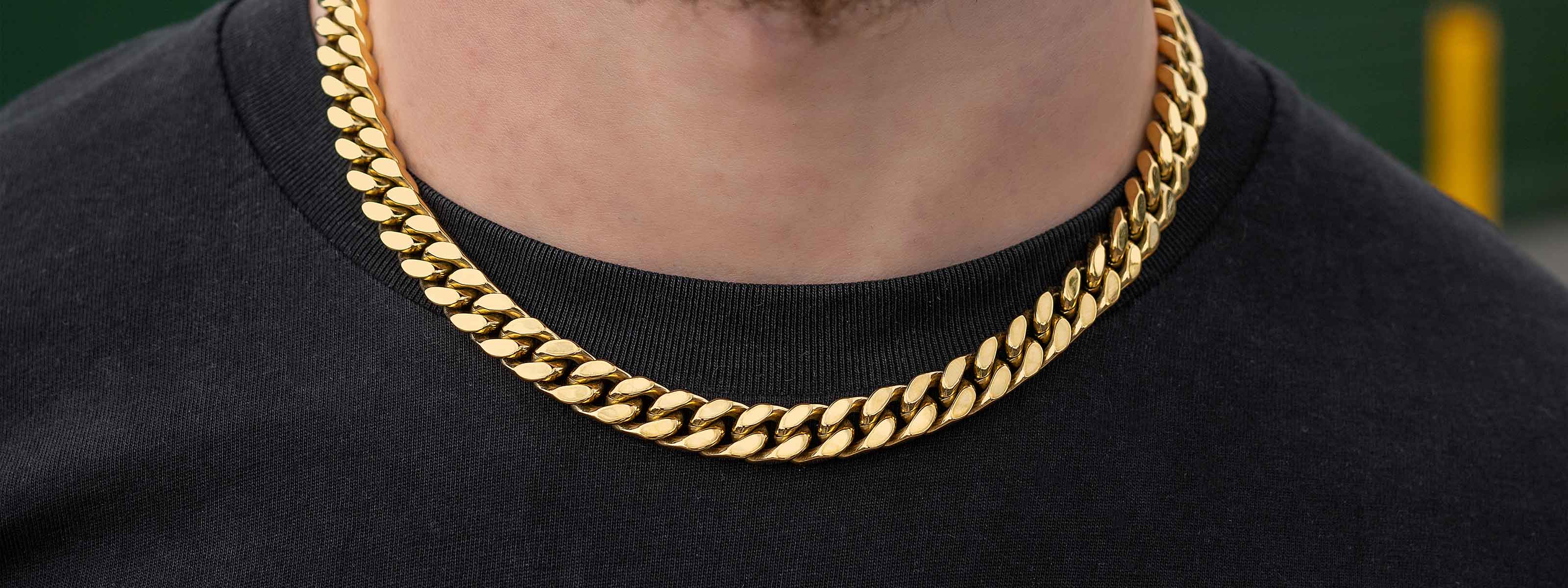 A close-up of a person wearing a thick gold Cuban link chain necklace over a black t-shirt. The persons neck and part of the chin are visible.