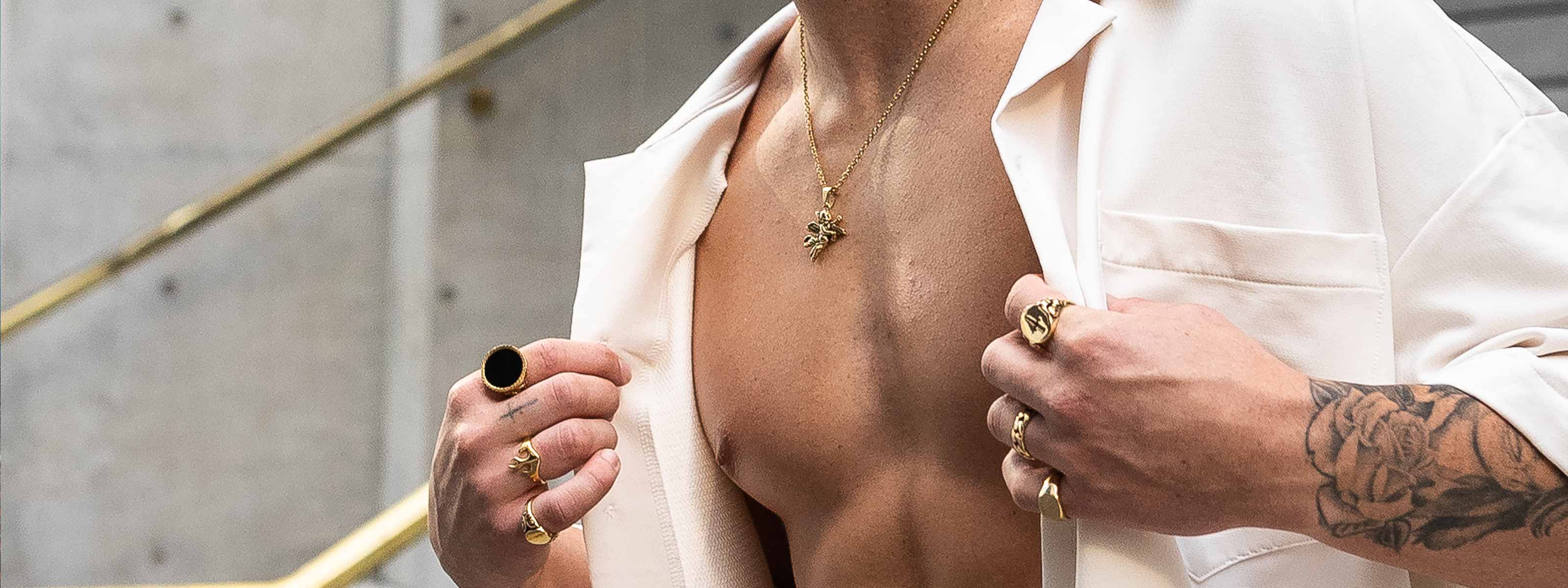 A person wearing an open white shirt displays layered gold necklaces, several gold rings, and has a tattooed arm. The background shows a modern setting with concrete walls and brass railings.