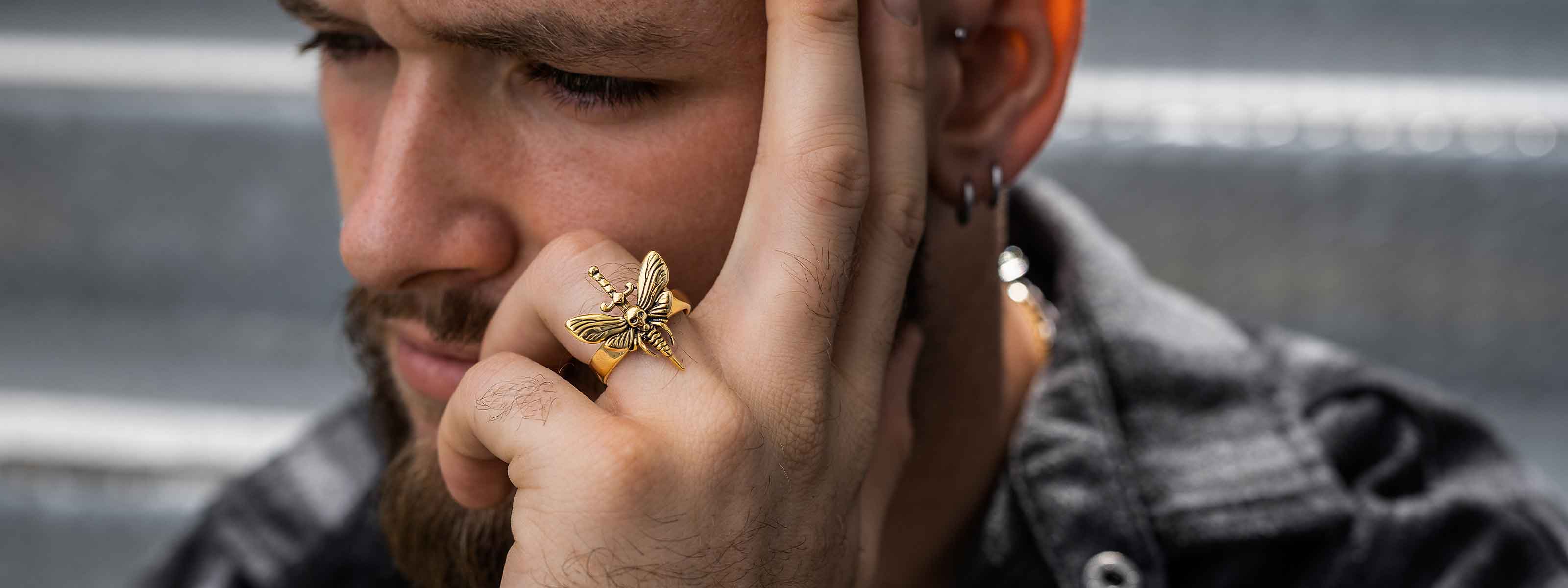 A man with a beard rests his chin on his hand, wearing a large gold butterfly ring and multiple earrings, with a blurred outdoor background.