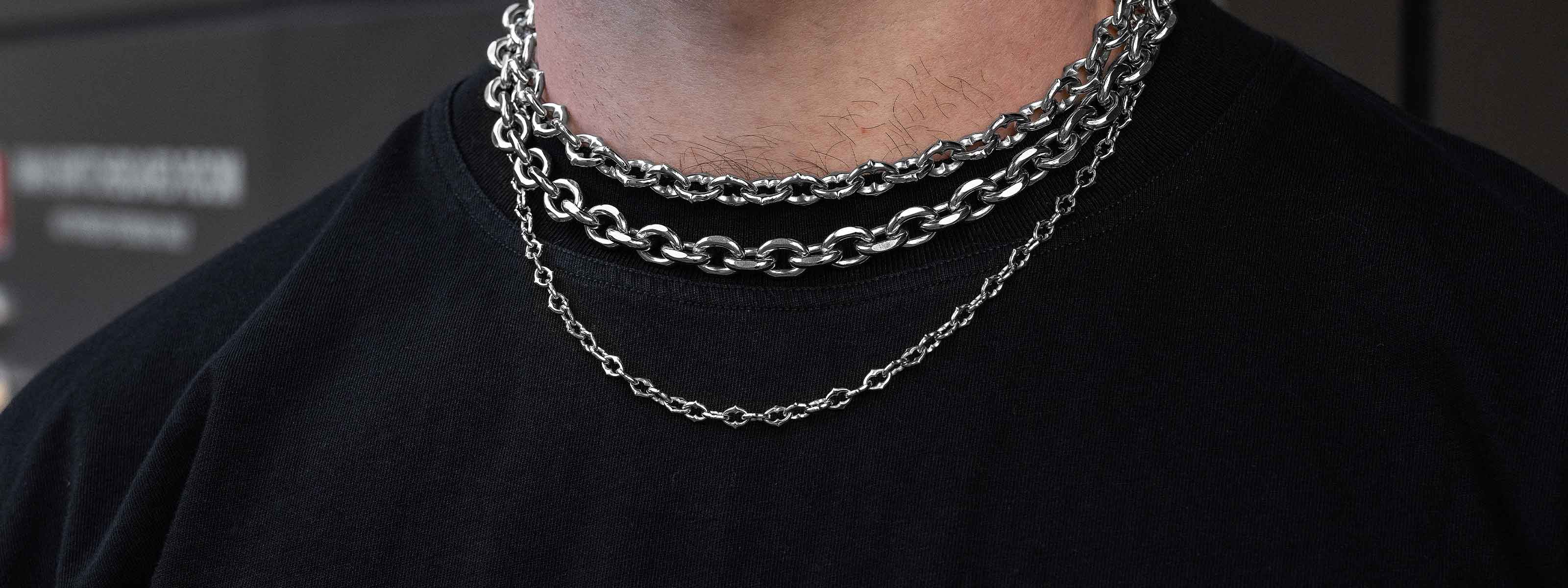 A person wearing a black shirt and three silver chains of varying thicknesses around their neck, with two chunky chains stacked above a thinner chain.