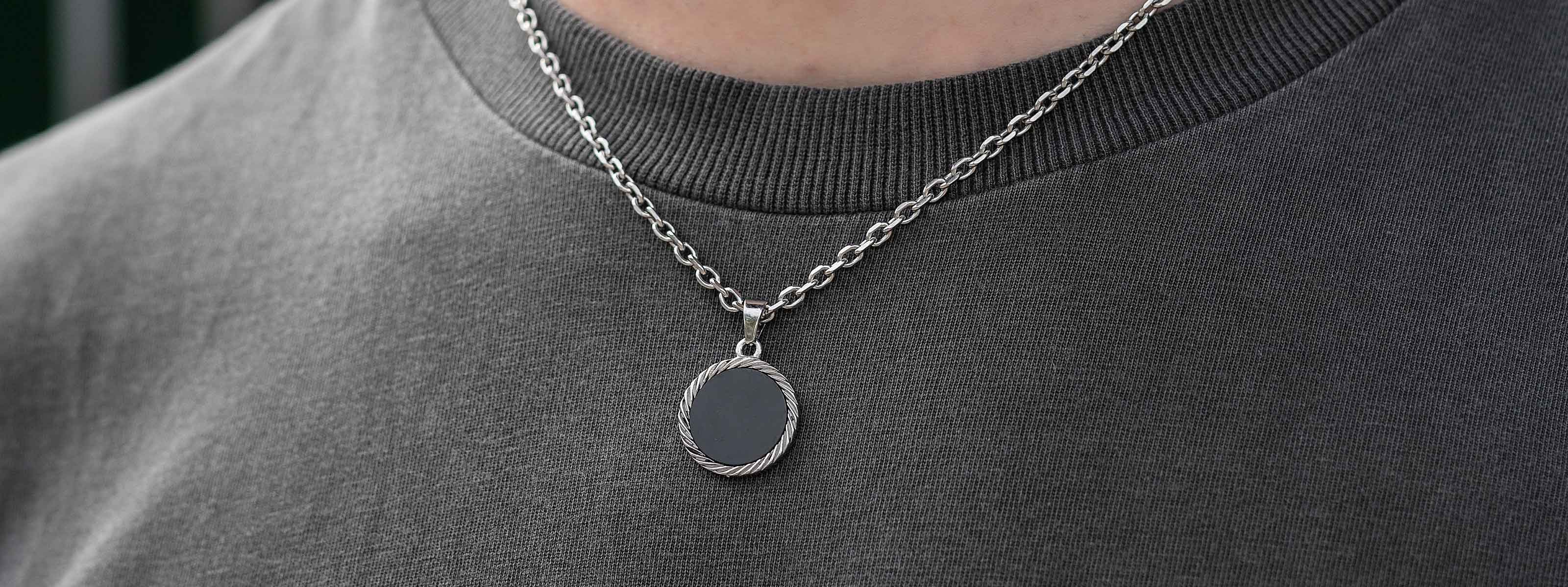 A close-up of a person wearing a grey shirt and a silver twisted chain necklace with a round, dark pendant.
