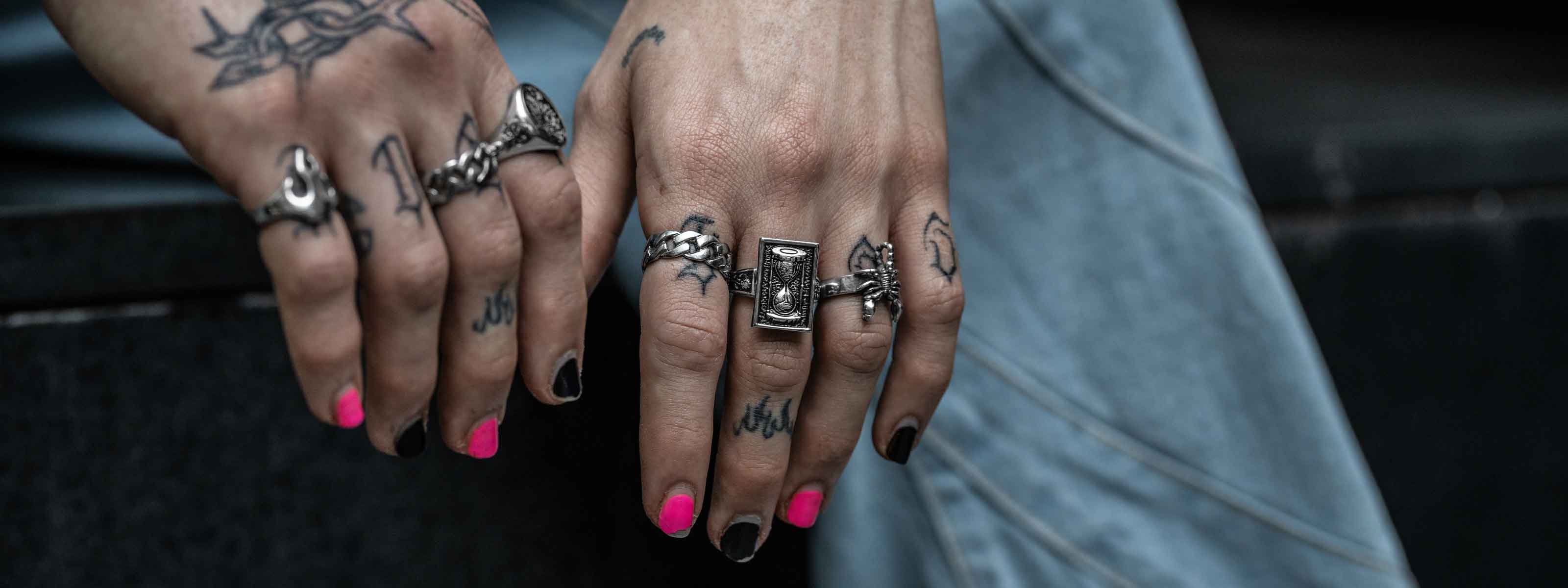 Close-up of two tattooed hands with multiple silver rings and black and pink nail polish, resting on denim-clad legs.