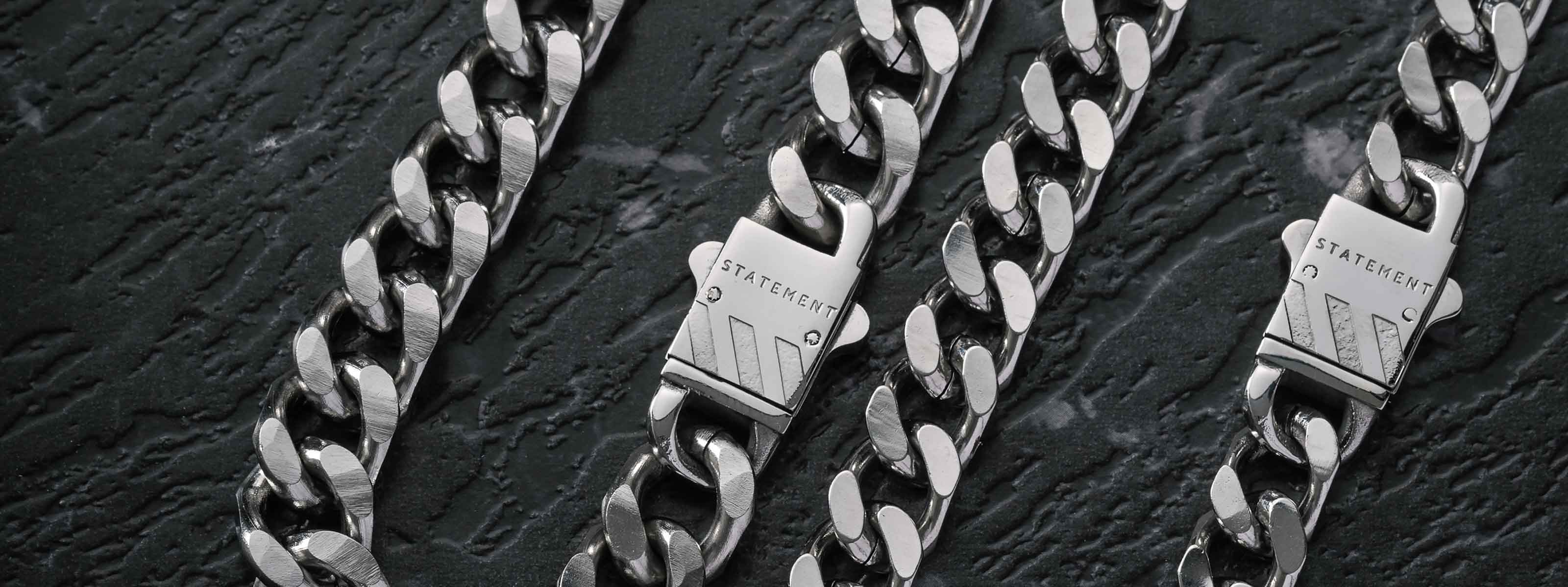 Three silver chain necklaces with rectangular clasps engraved with the word STATEMENT are arranged on a dark, textured stone surface.