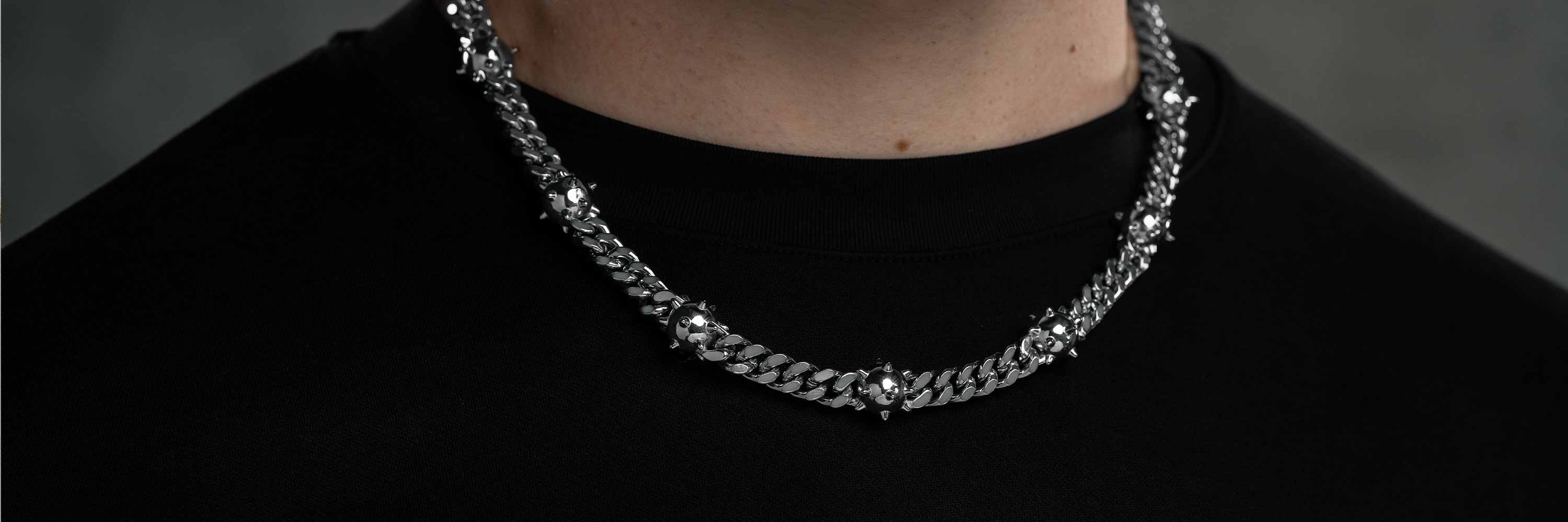 A close-up of a person wearing a thick silver chain necklace with decorative details, over a black crew-neck shirt, against a dark, blurred background.