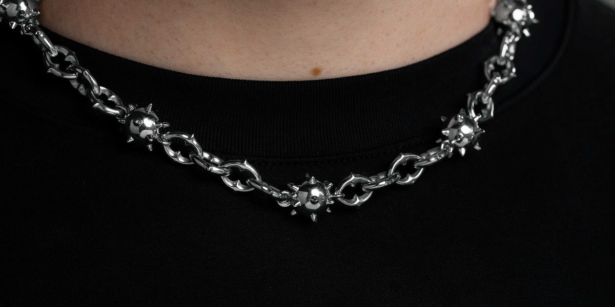 A close-up of a person wearing a thick silver chain necklace with decorative details, over a black crew-neck shirt, against a dark, blurred background.