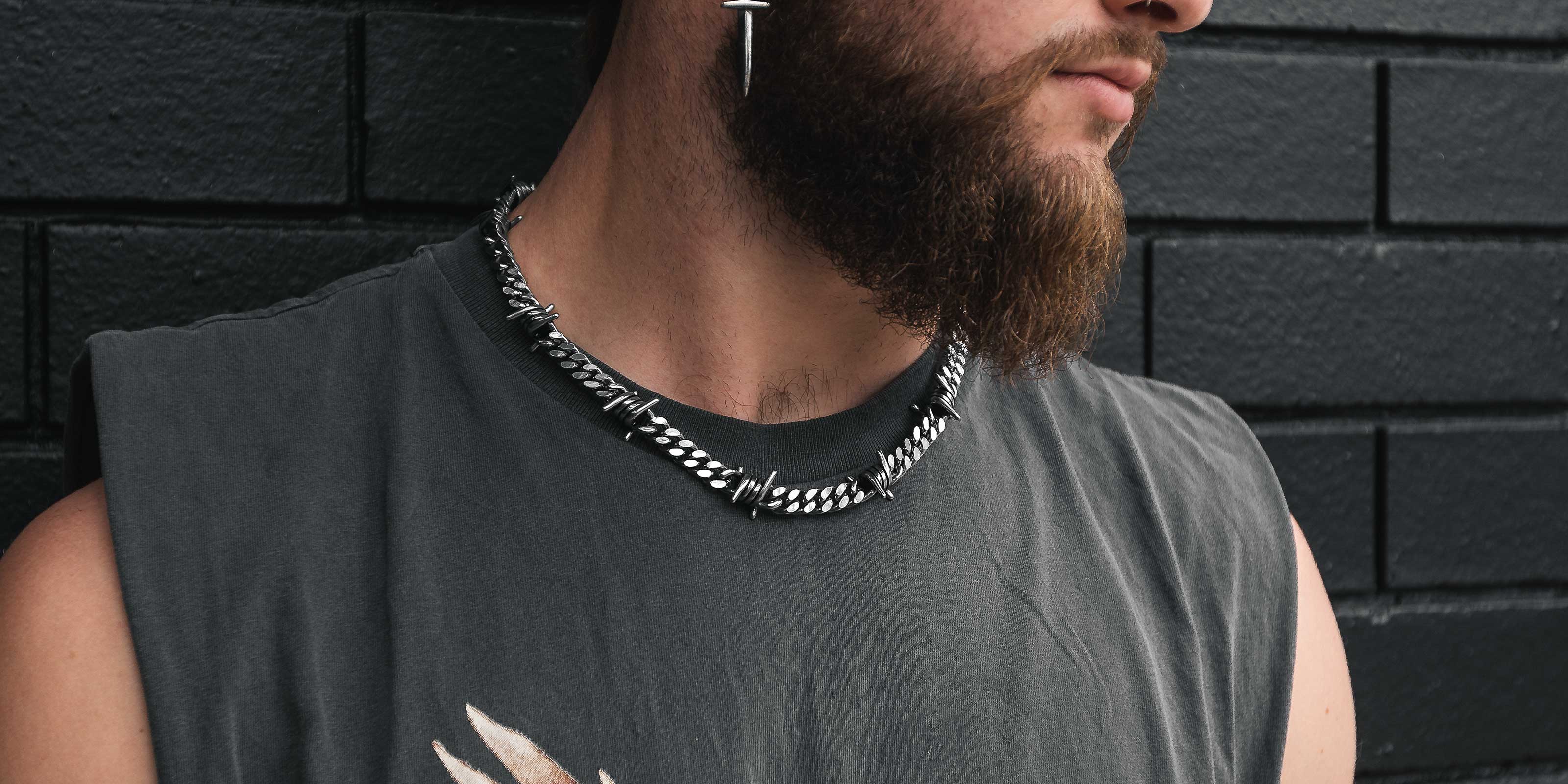 A person wearing a shiny silver chain necklace with textured links over a plain black shirt. The background is dark and out of focus.