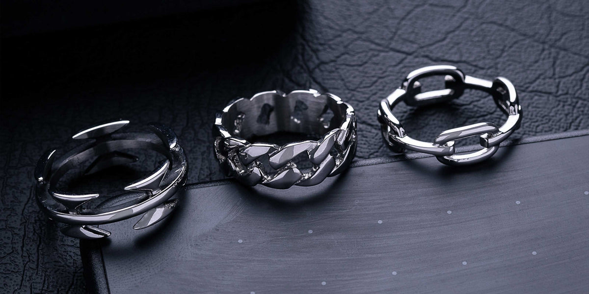 A close-up of a hand with four silver rings on the fingers, each with a unique design, held in a fist against a blurred brown background.