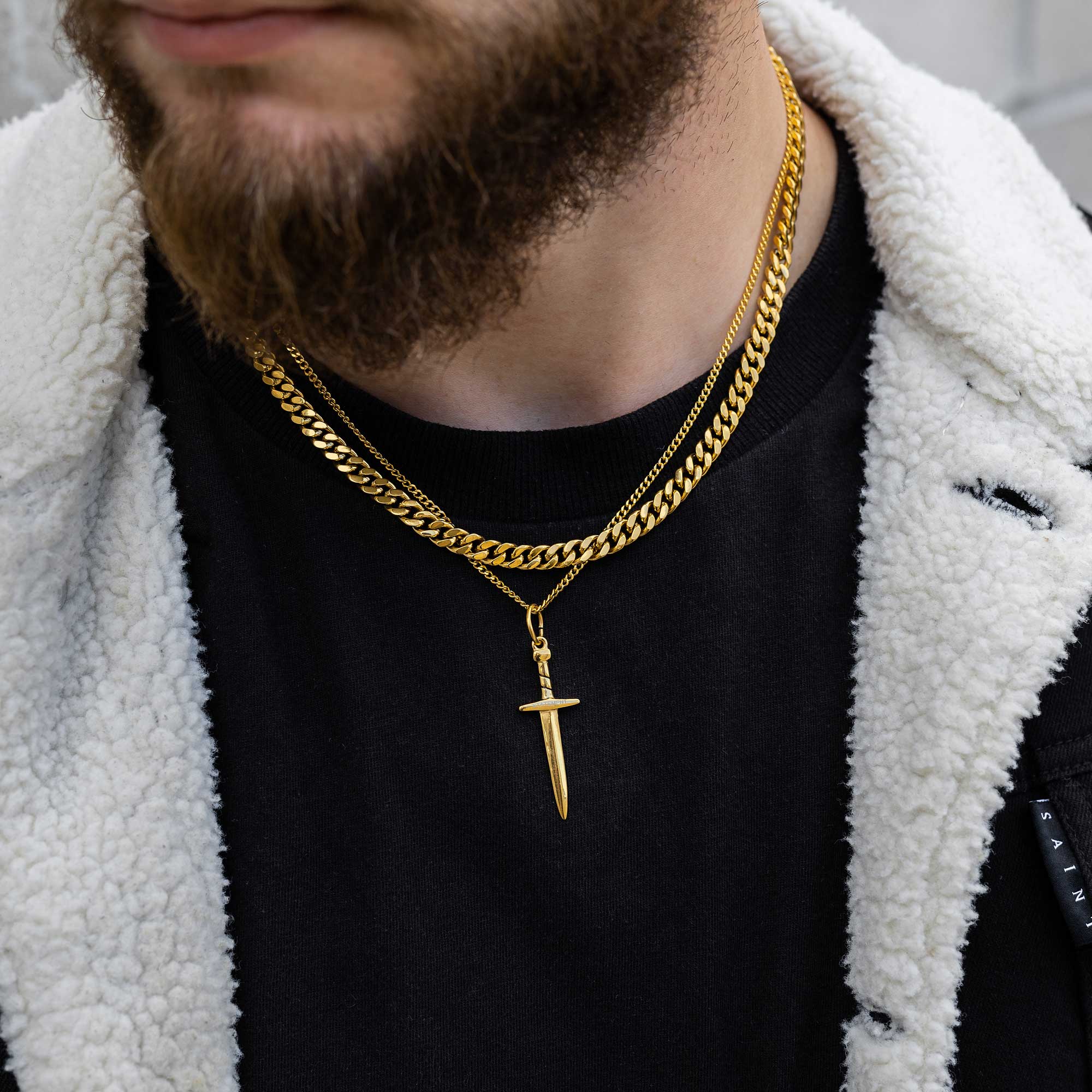 A man with a beard wears a black shirt, a white sherpa jacket, and two gold chains, one with a sword pendant and the other a thick curb link chain.