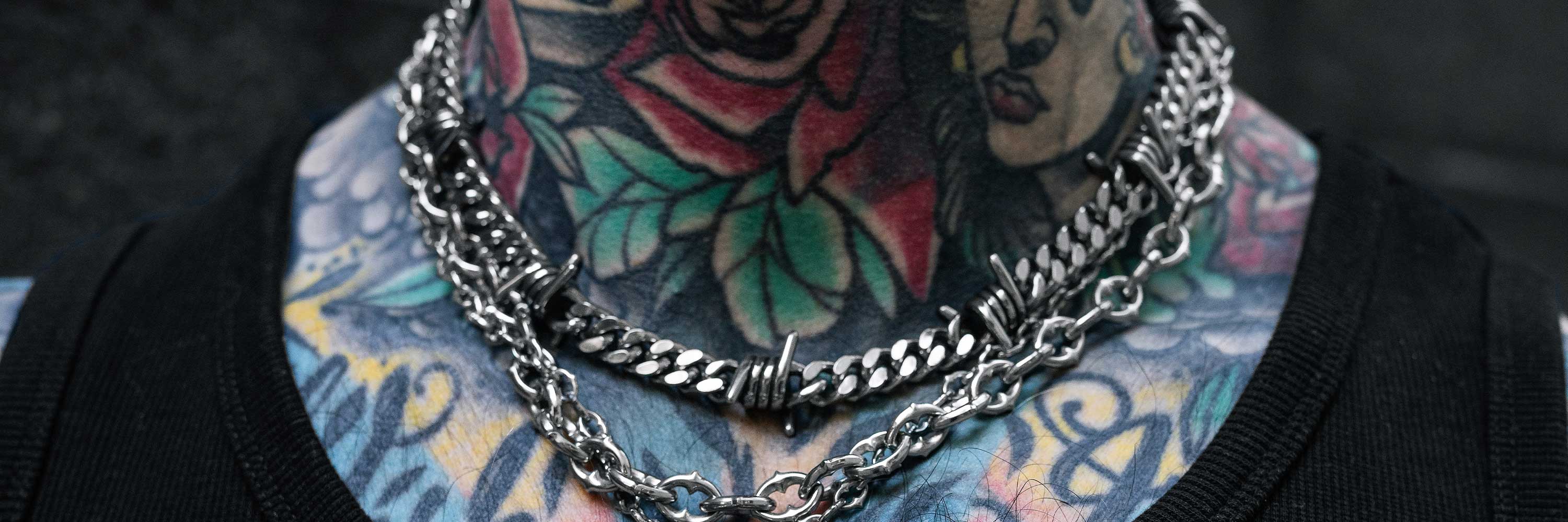 A close-up of a tattooed neck and upper chest wearing layered silver chains and barbed-wire style necklaces. The colorful tattoos include floral and portrait designs.