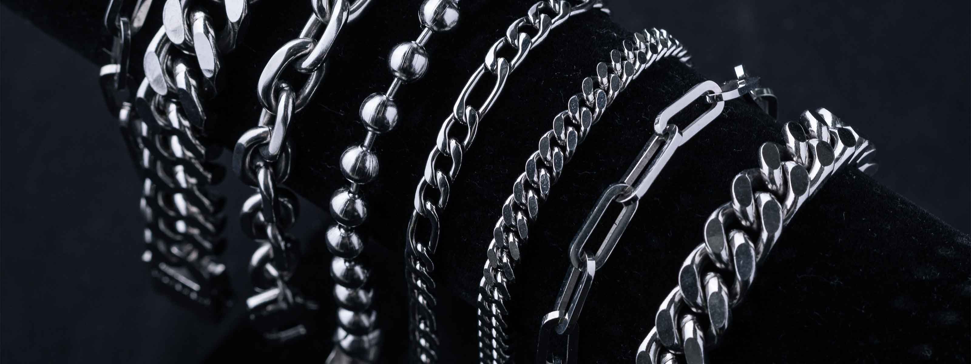 A display of various silver chain bracelets and necklaces with different link styles arranged on a black velvet stand against a dark background.