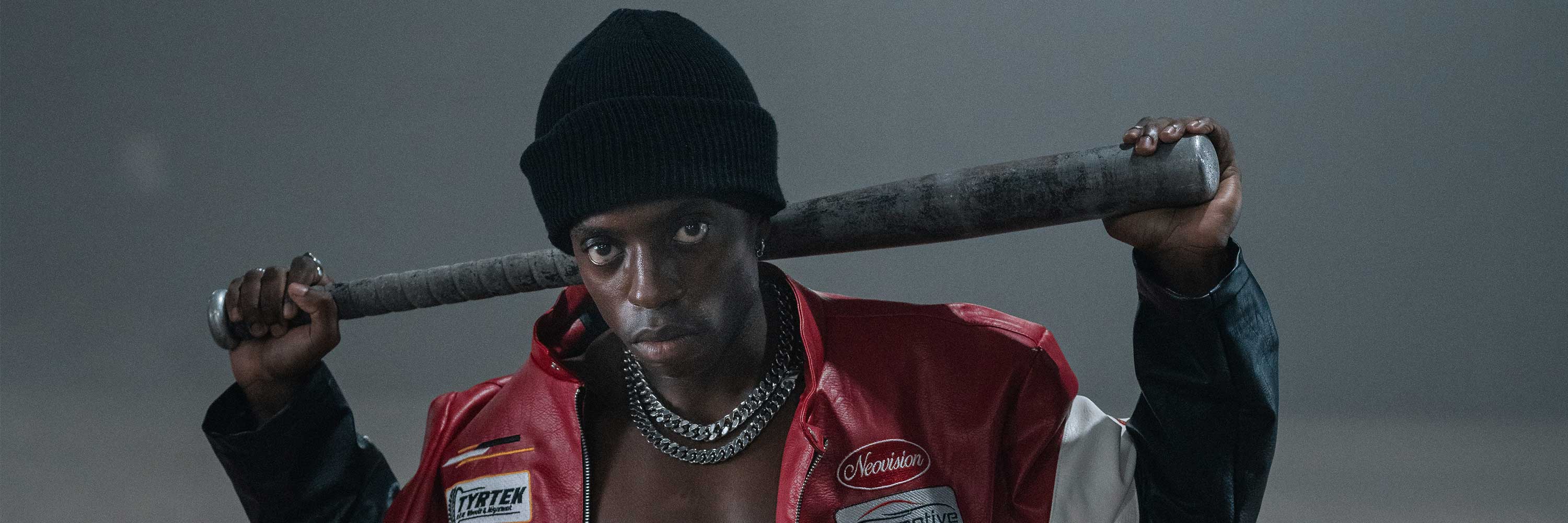 A serious-looking person in a red and black jacket and black beanie holds a baseball bat across their shoulders, wearing chunky silver chains against a dark, foggy background.
