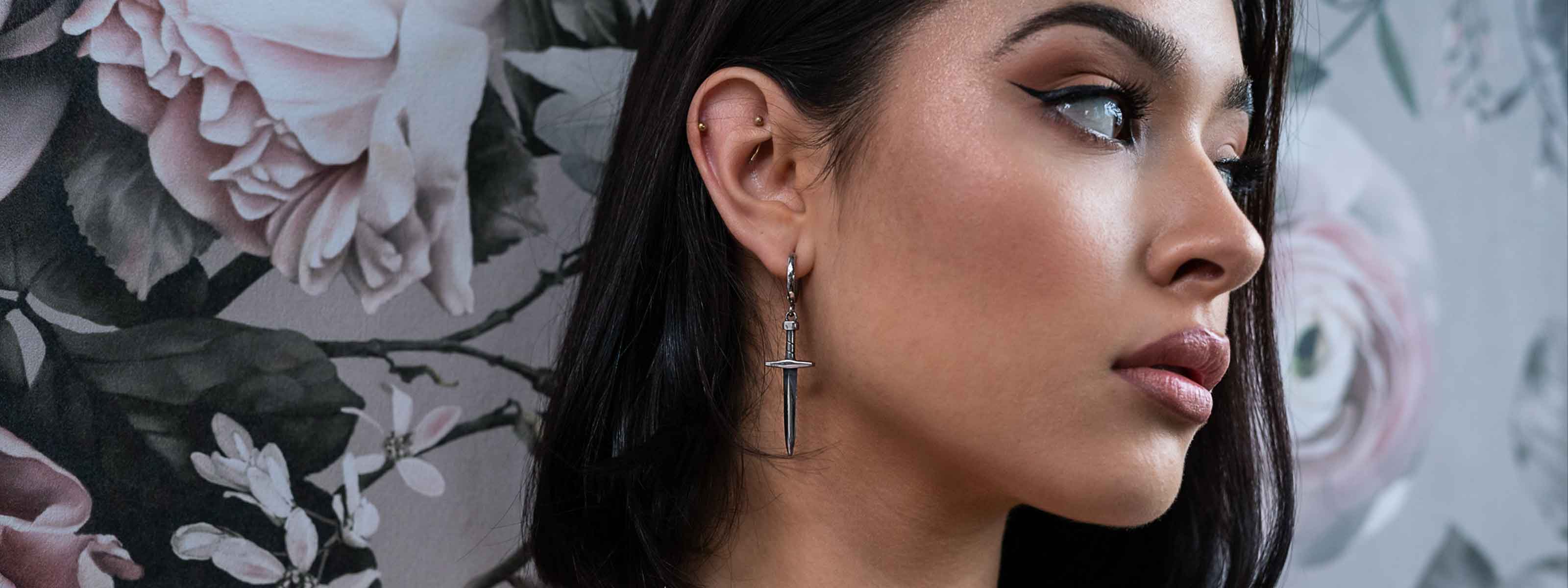A woman with dark hair and winged eyeliner wears a sword-shaped earring, posing in profile against a floral wallpaper background.