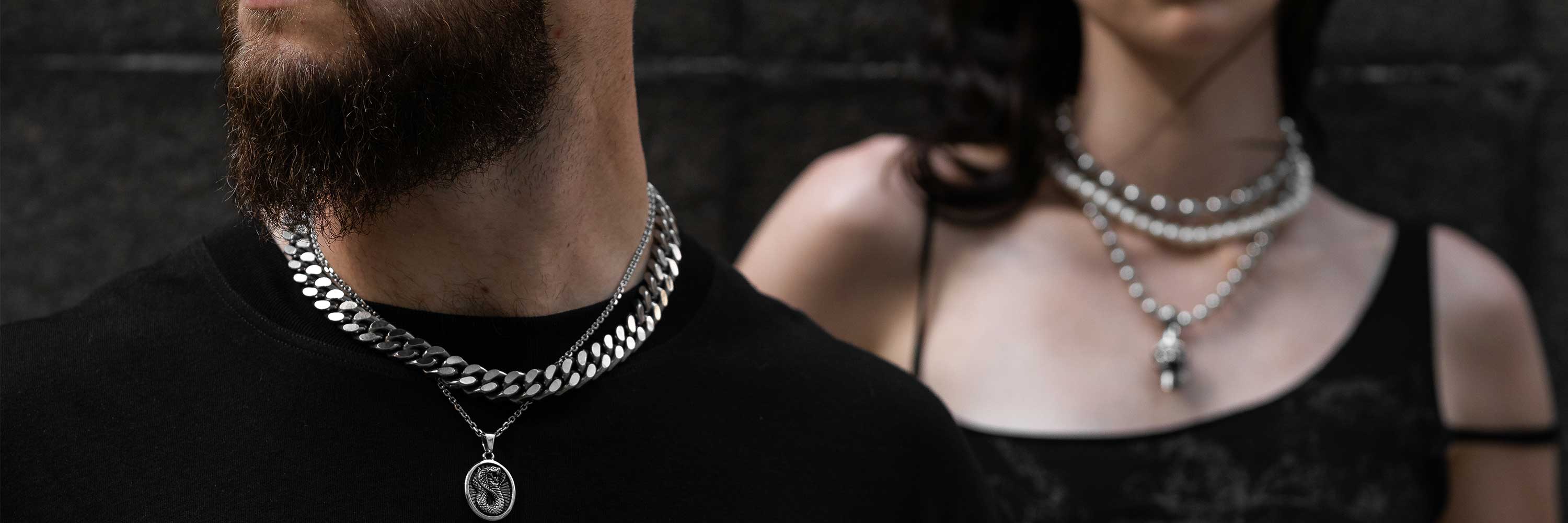 Close-up of a man and woman wearing layered silver chain necklaces; the man also has a pendant and a beard, while the woman’s necklaces include a pendant and pearls. Both are dressed in black.