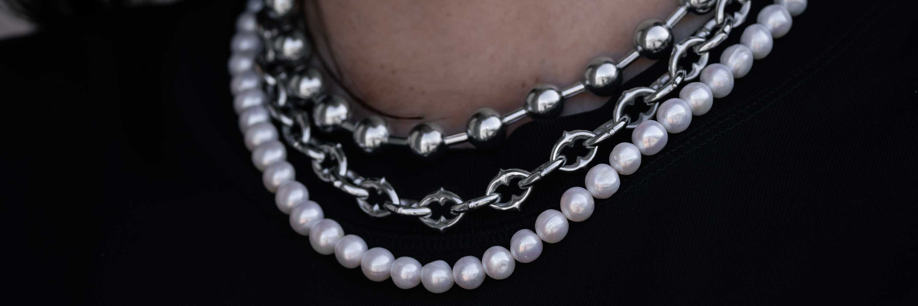 Close-up of a person wearing layered necklaces: one with white pearls and two with chunky silver chains, all against a black top.