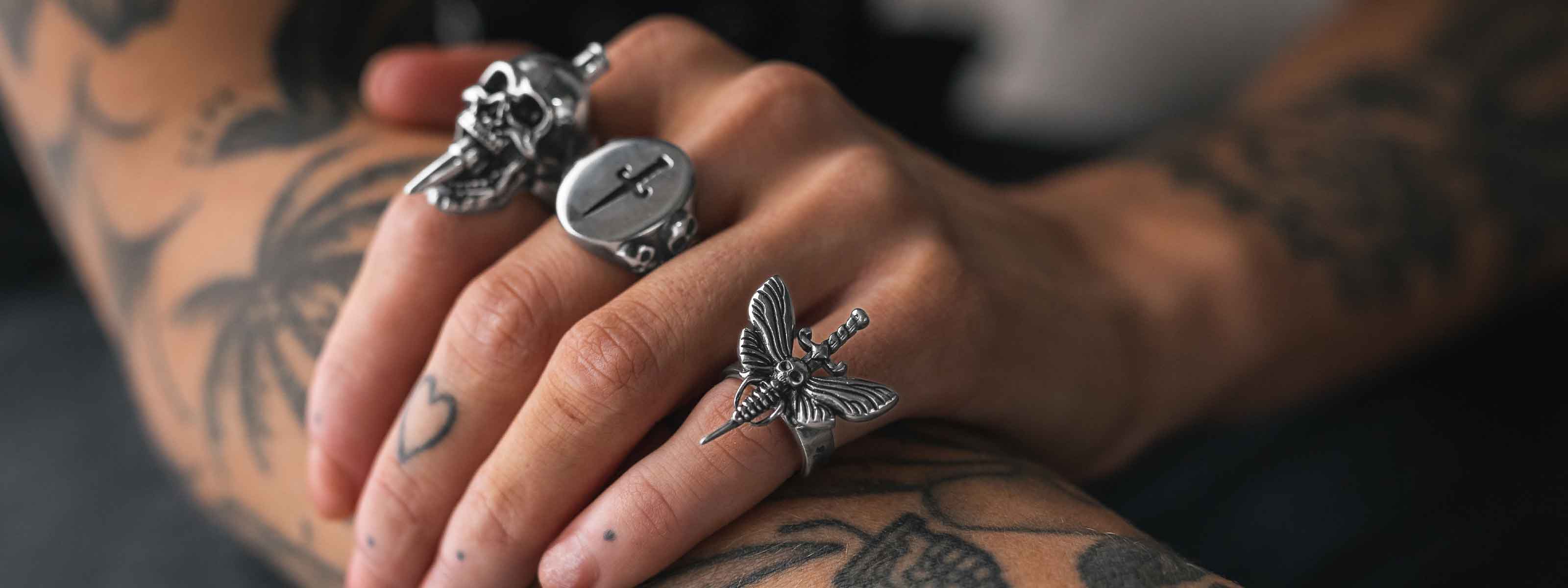 A close-up of tattooed hands wearing bold silver rings, including designs of a skull, a button, and a butterfly with a sword. Small heart and dot tattoos are visible on the fingers.