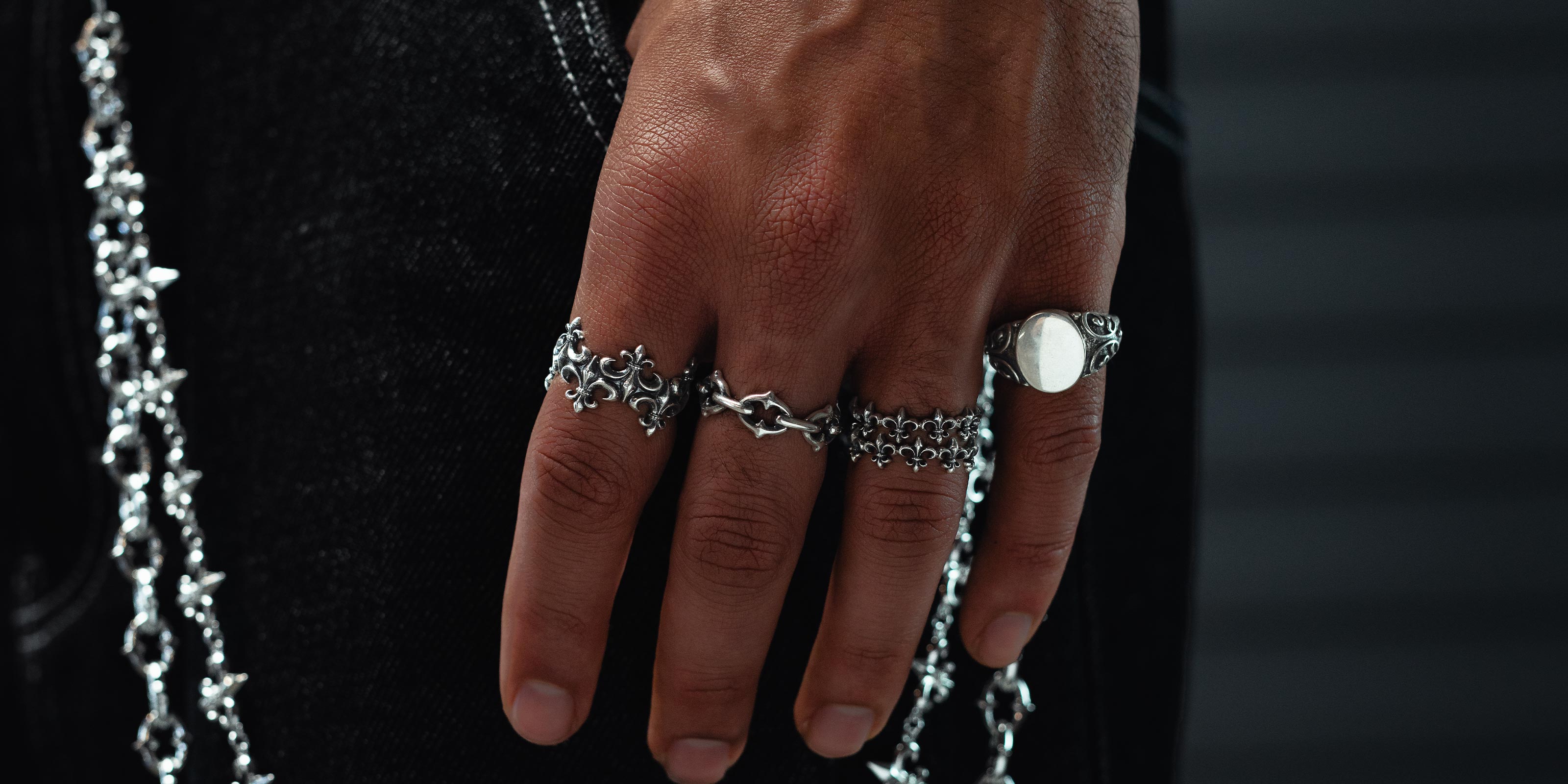 A close-up of tattooed hands wearing bold silver rings, including designs of a skull, a button, and a butterfly with a sword. Small heart and dot tattoos are visible on the fingers.