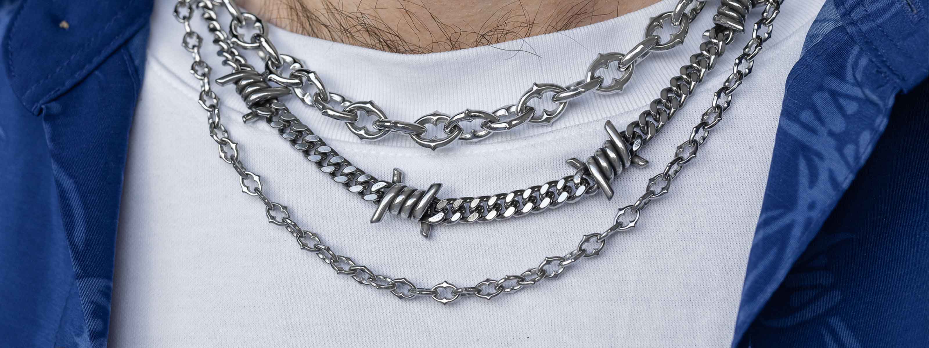 A person wearing a white shirt and blue outerwear has layered silver necklaces, including a barbed wire chain, around their neck.
