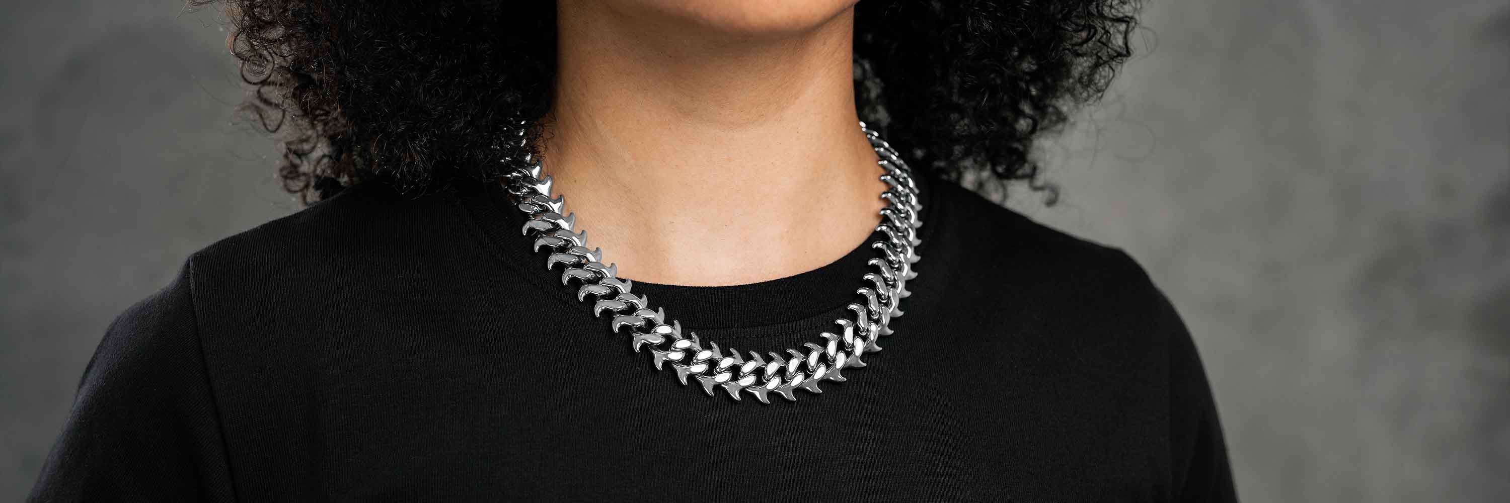 A person with curly dark hair wearing a black top and two thick, silver chain necklaces against a neutral grey background.