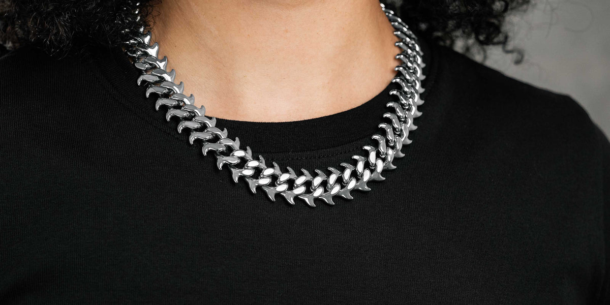 A person with curly dark hair wearing a black top and two thick, silver chain necklaces against a neutral grey background.