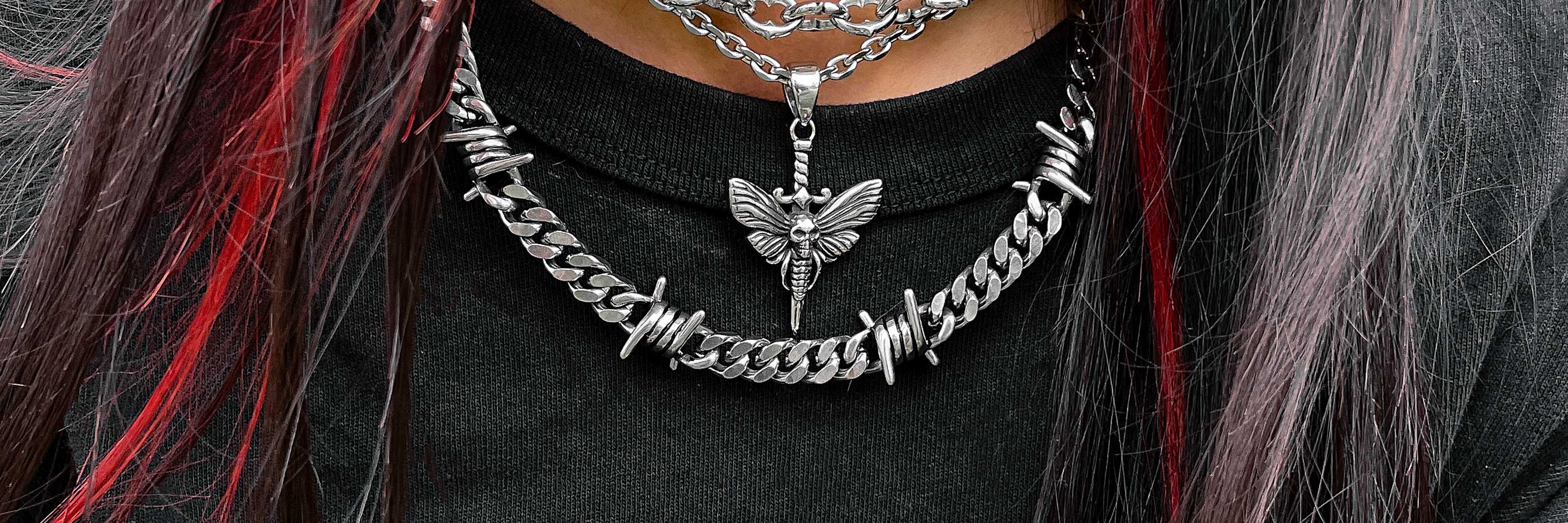 Close-up of a person wearing layered silver necklaces, including a barbed wire chain and a pendant shaped like a moth, with long dark hair featuring red highlights.