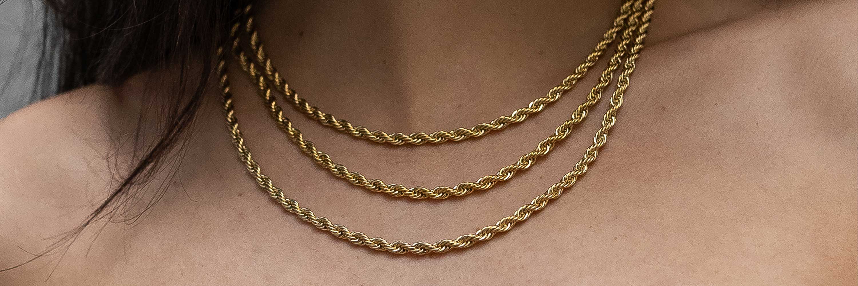 Close-up of a person’s neck wearing three layered gold rope chain necklaces, each of a different length, against bare skin.