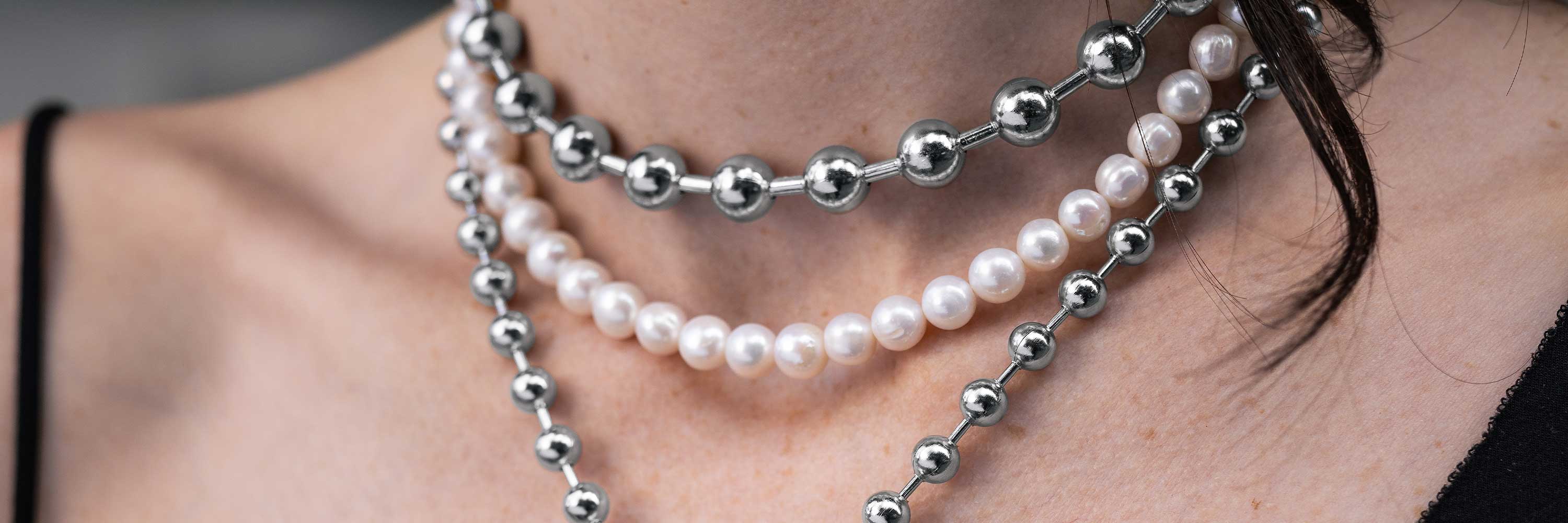 Close-up of a person wearing layered necklaces, including silver metallic beads and white pearl beads, over bare skin and a black spaghetti strap top.
