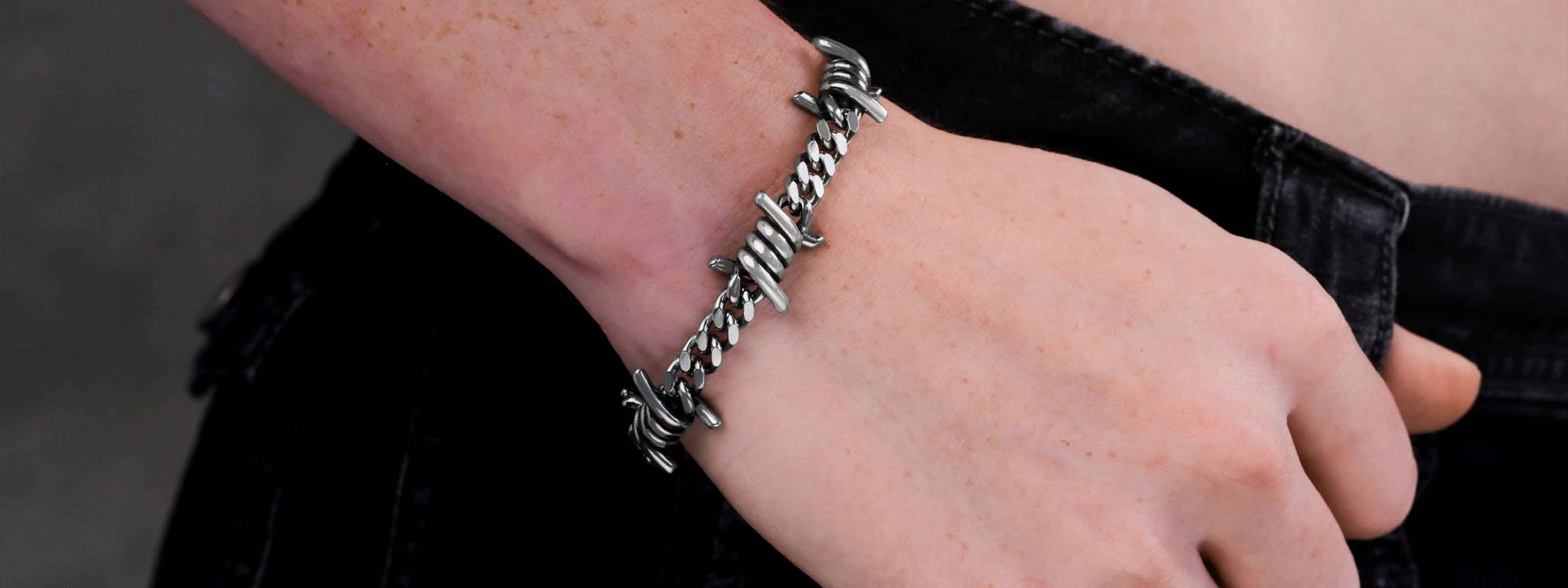 A person wearing a silver bracelet designed to look like barbed wire rests their hand near the pocket of dark jeans.
