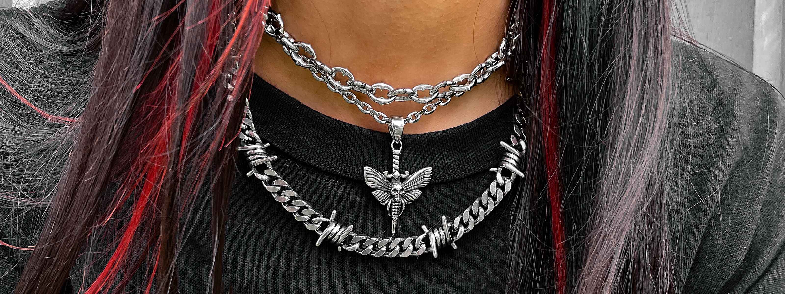 Close-up of a person wearing two layered silver chain necklaces, one with a butterfly pendant and the other with barbed wire details, over a black shirt. The person has long dark hair with red highlights.