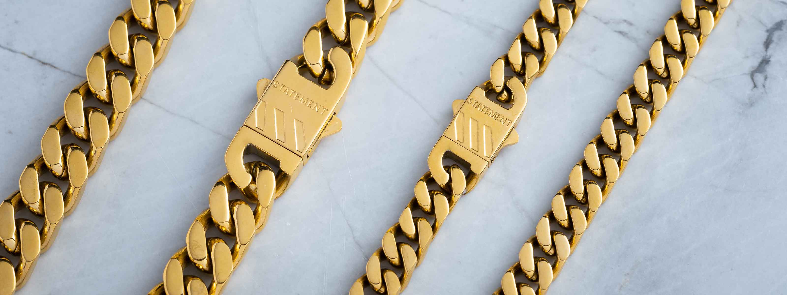 Four gold chain necklaces with clasps are displayed on a light marble surface. The chains vary in thickness, and the clasps are engraved with the word CUBANLINKS.