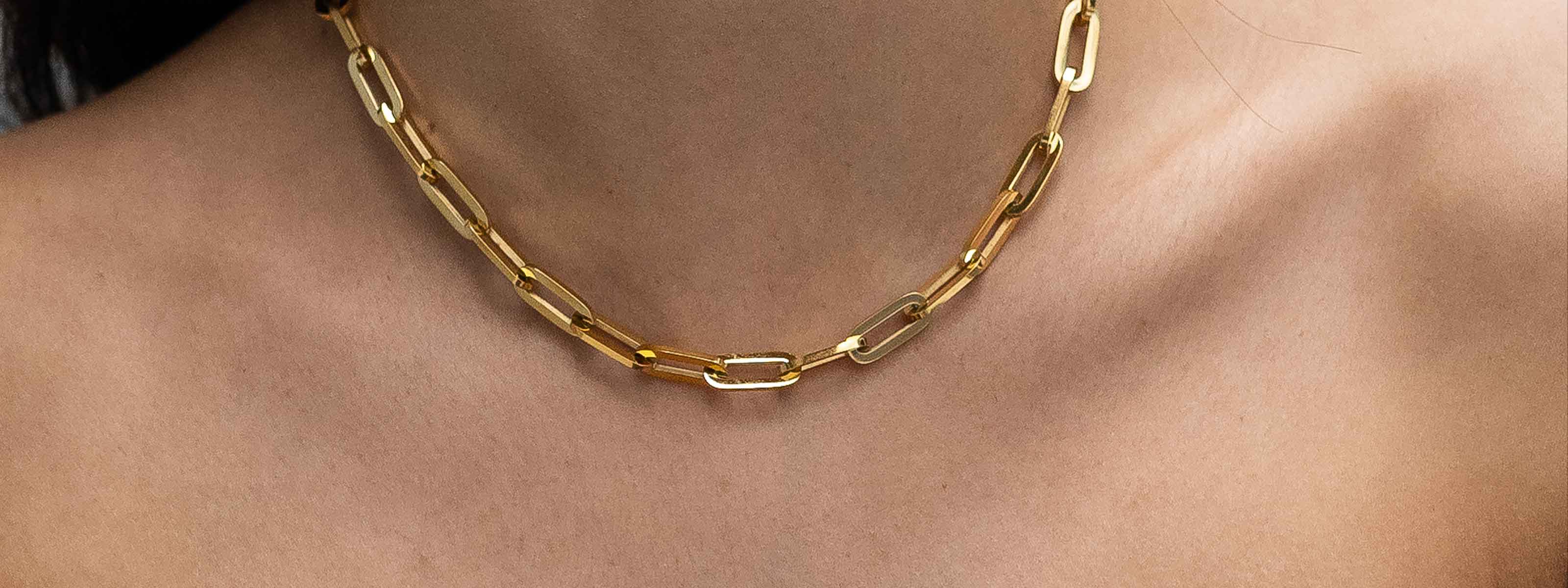 A close-up of a womans neck wearing a gold chain necklace with elongated, rectangular links against bare skin.