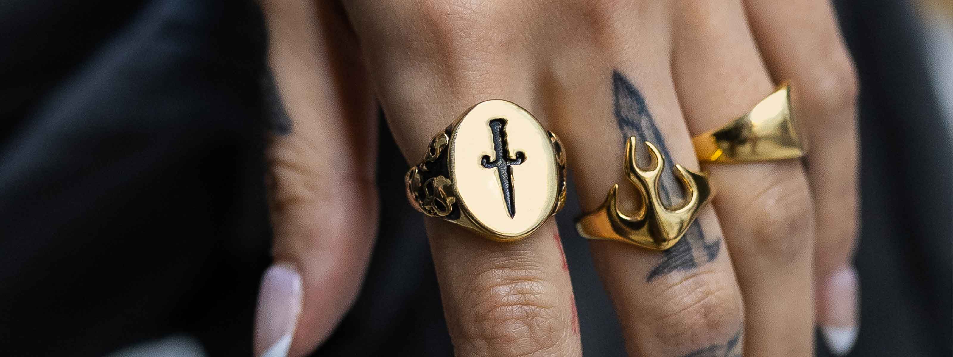 A close-up of a hand with black nail polish wearing three gold rings, including one with a black sword design and another shaped like a trident, with a black tattoo visible on the finger.