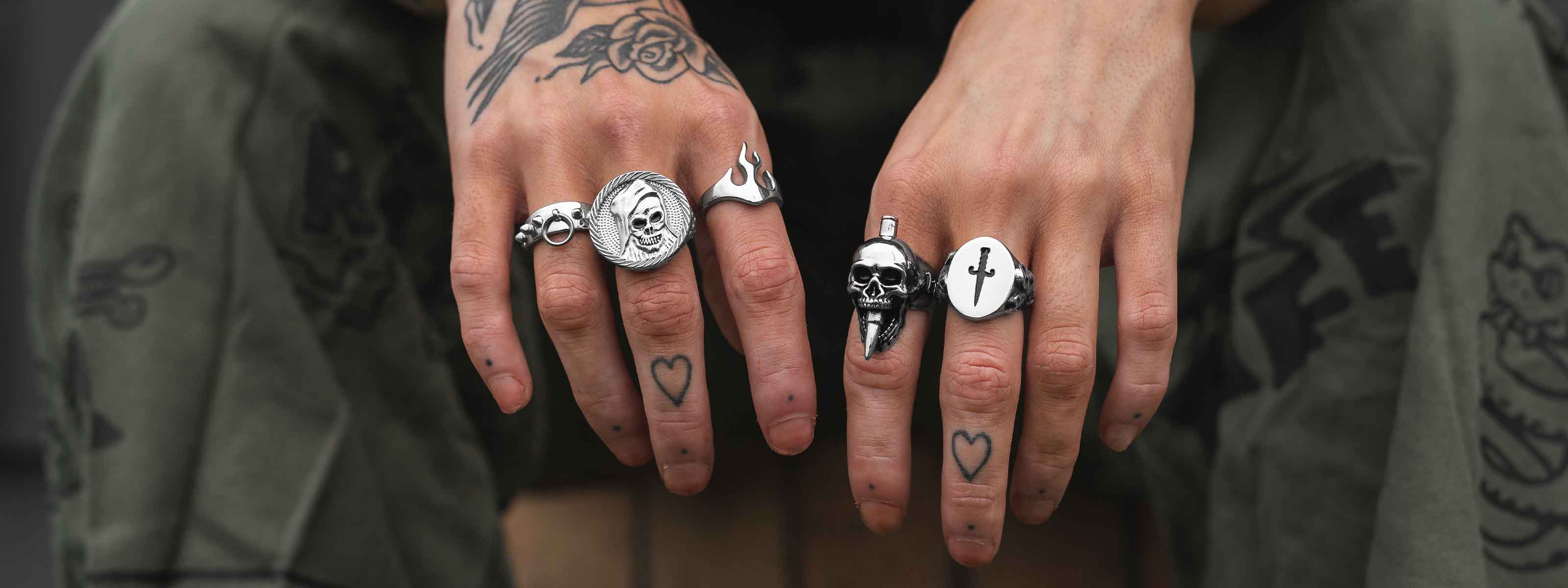 A close-up of tattooed hands wearing several silver rings with skull, flame, and cross designs. Heart tattoos are visible on the fingers, and the person wears camo-patterned pants.