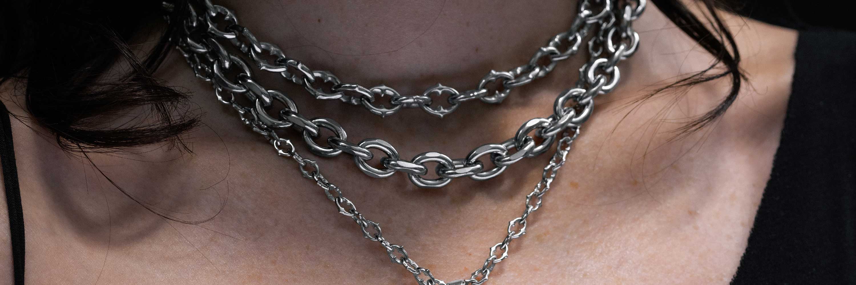 A close-up of a person wearing three layered silver chain necklaces around their neck, with dark hair visible and a black top.