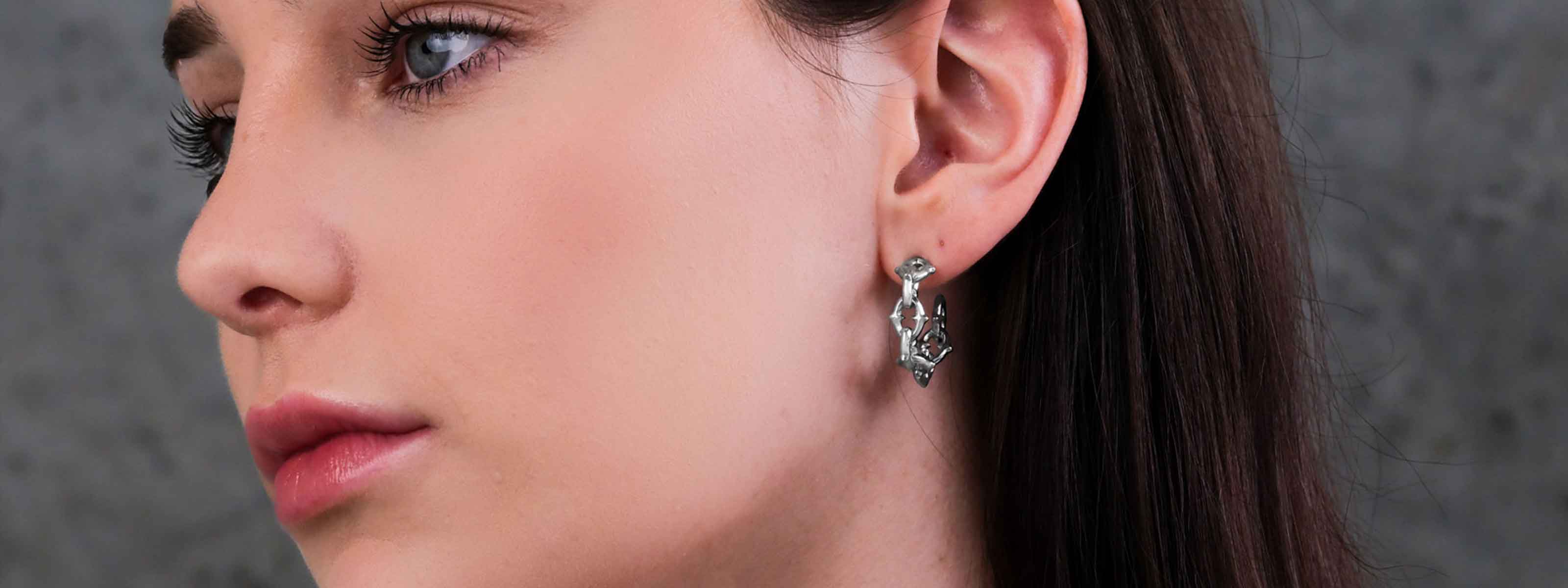 Close-up of a woman with fair skin and dark hair, wearing intricate silver earrings shaped with geometric patterns, against a blurred gray background.